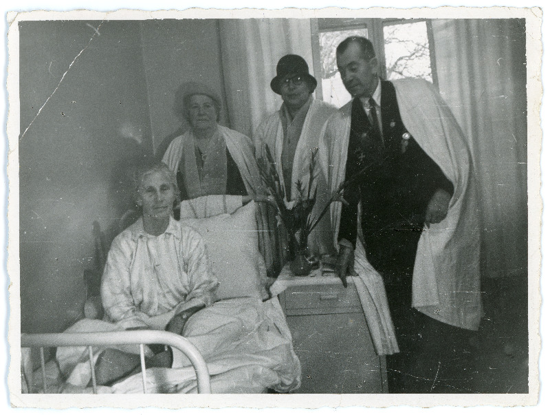 Tundmatu naisterahvas haiglavoodil, kõrval kolm külastajat kitlites, öökapil lilled. Pöördel venekeelne loetamatu tekst