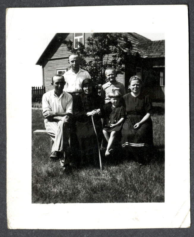 Osvald Asi perekonnafoto: mees, musta rõivastatud vanem naine kepiga, tüdruklaps ja noor naisterahvas istuvad pingil, meesterahvas ja poisslaps seisavad nende taga. Taustal elumaja