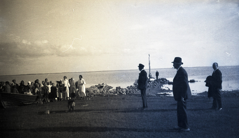 Vaade rannale, paremal seismas kolm ülikondades meest, vasakul grupp [kohalikke?] inimesi