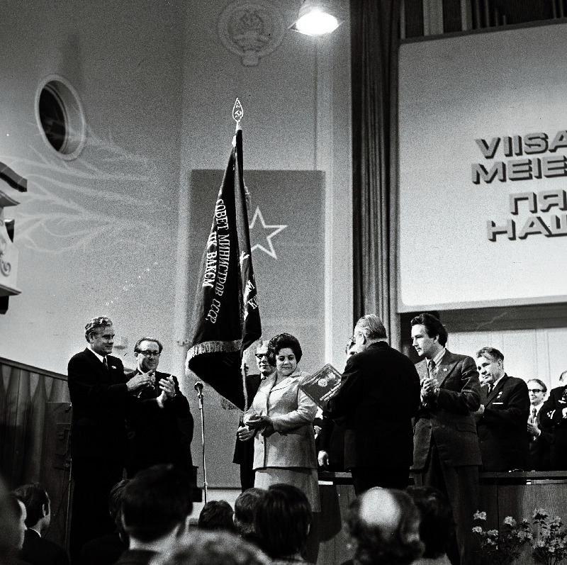 1974. a. üleliidulise sotsialistliku võistluse tulemuste põhjal Tallinnale määratud NLKP Keskkomitee, NSV Liidu Ministrite Nõukogu, ÜAÜKN-i ja ÜLKNÜ Keskkomitee rändpunalippu hoiab sotsialistliku töö kangelane, Tallinna linna aukodanik A. Valdov. Vasakult 1. EKP Tallinna Linnakomitee esimene sekretär N. Johanson, 2. Tallinna TSN Täitevkomitee esimees J. Kallion, 6. EKP Keskkomitee esimene sekretär J. Käbin.