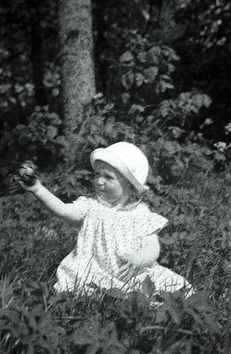 Väike täpulises kleidis ning kübaras tüdruk istub metsas, rohu peal.