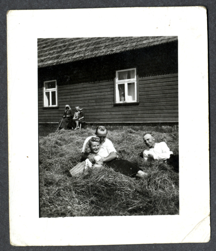 Osvald Asi, tundmatu naine ja mees lamavad elumaja ees heinakuhjas. Maja seina ääres istub vanem naisterahvas koos kahe väikese lapsega