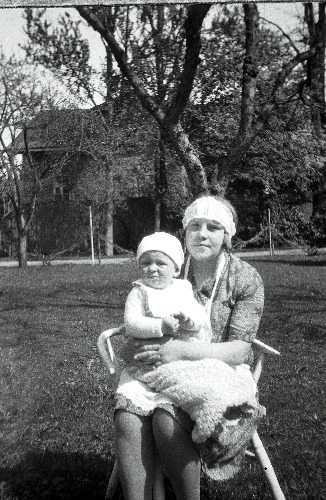 Suures aias istub toolil naine, väike laps süles. Tagapool näha hooneid.