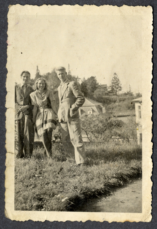 Neiu ja kaks noormeest seisavad haljasalal. Selja taga paistavad majad