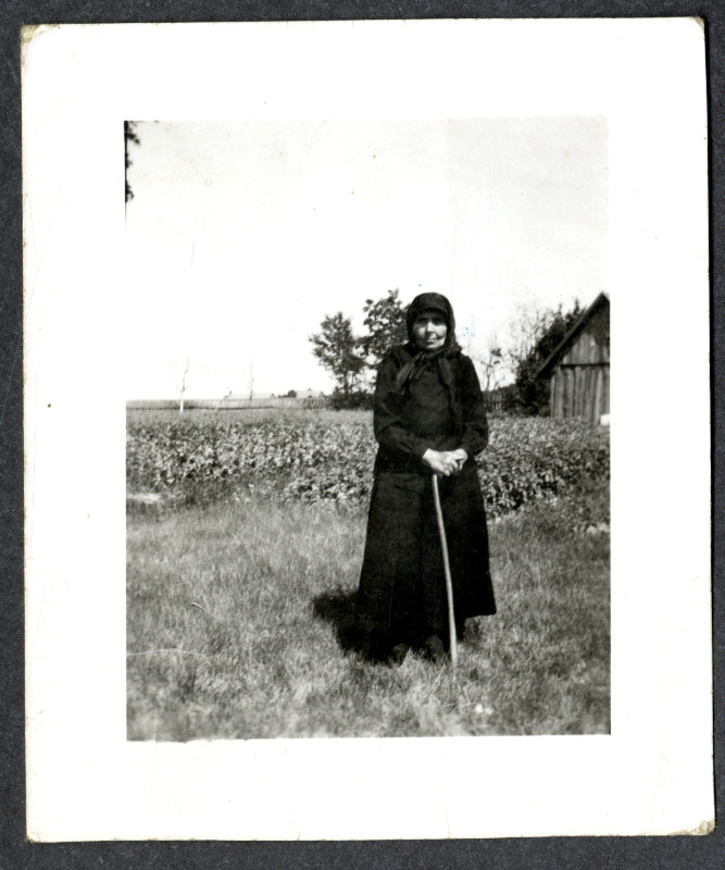 Musta rõivastatud pearätikuga vanem naine seisab kepile toetudes peenra või põllu ääres. Eemalt paistab taluhoone nurk, puud ja aed