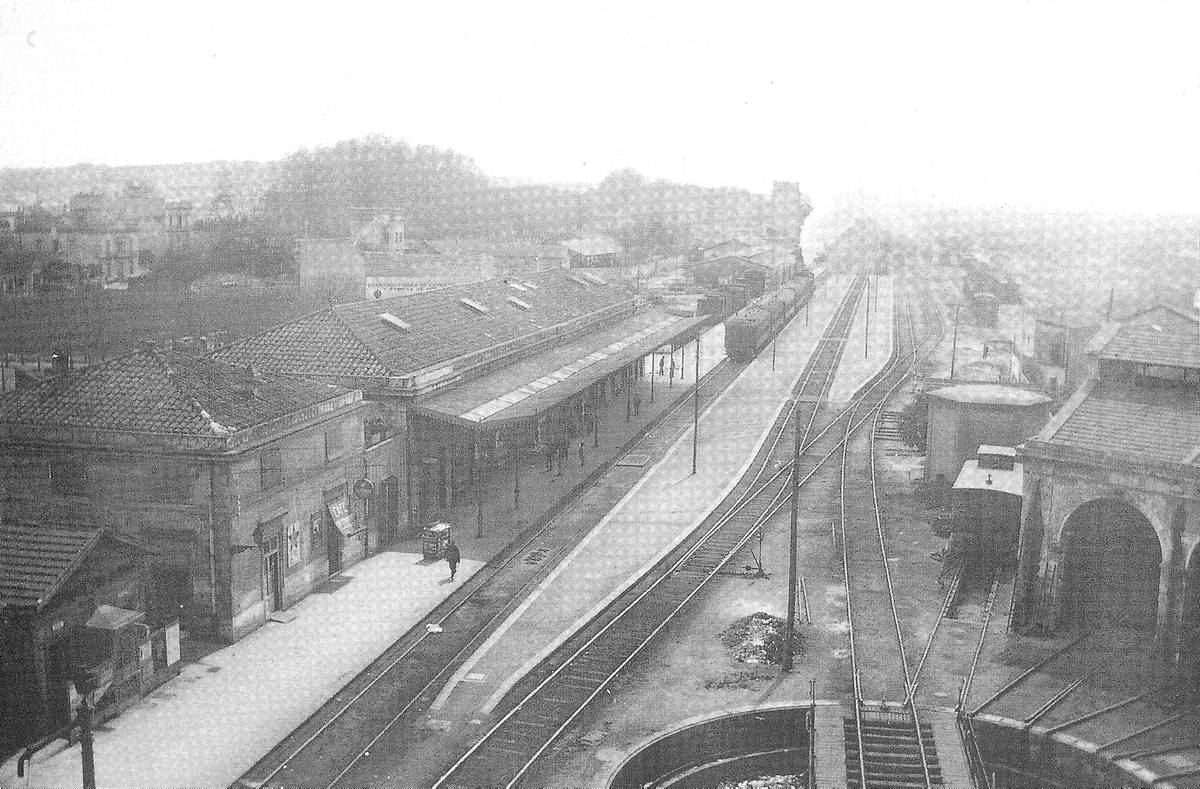 [Girona train station] - View from a high point of the train station of Girona with a convoy arriving. In the background on the left, the train station of Sant Feliu and the road to Barcelona. On the right, part of the railway company’s facilities and warehouses