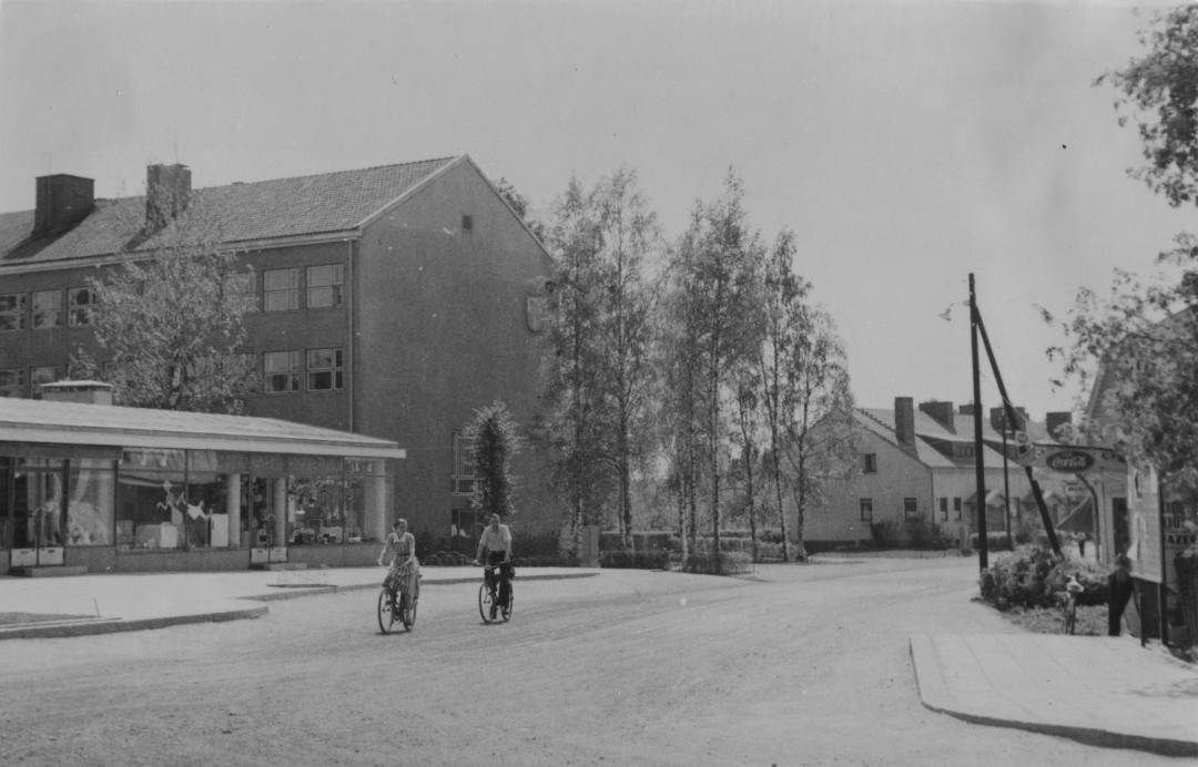 Katunäkymä Aleksis Kiven tien varrelta 1950-luvun jälkipuoliskolta. Vasemmalla vuonna 1957 valmistuneen Suomen Työväen Säästöpankin (STS) matala liikesiipi, jonka takana Keravan kauppalantalo. Kauppalantalon päädyssä Keravan ensimmäinen kuntavaakuna. Kauppalantalon takana näkyy vuonna 1952 valmistuneet kauppalan virkailijoiden lamellitalo (ns. haitaritalo). Tien oikealla puolen maalarimestari Blommin talon kulmaa, jossa kuvaushetkellä toimi Kelloliike T.Soikkeli.  Päällystämättömällä tiellä pyöräilee pariskunta.
https://muistaja.finna.fi/keravan_museopalvelut/Record/kerava_mui.mui-15460