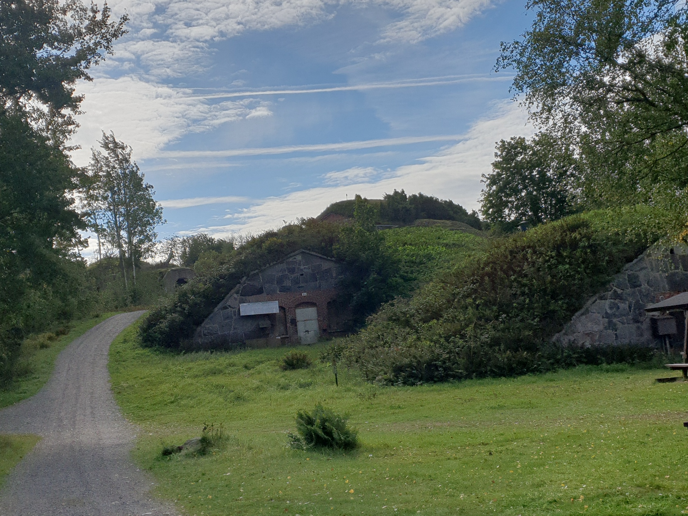 Näkymä Vallisaaren linnoituksilta saaren luoteisrannikolla. Maastoon on rakennettu kiviseinäisiä ruutivarastoja, jotka venäläiset rakensivat 1860-1870-luvuilla. Ne on katettu maakerroksella, ja ne saavat maaston kumpuilemaan. Hiekkainen kävelytie kiemurtelee kumpujen välistä. Vasemmalla näkyy holvikaarimainen betoninen sisäänkäynti varastoon. rephoto