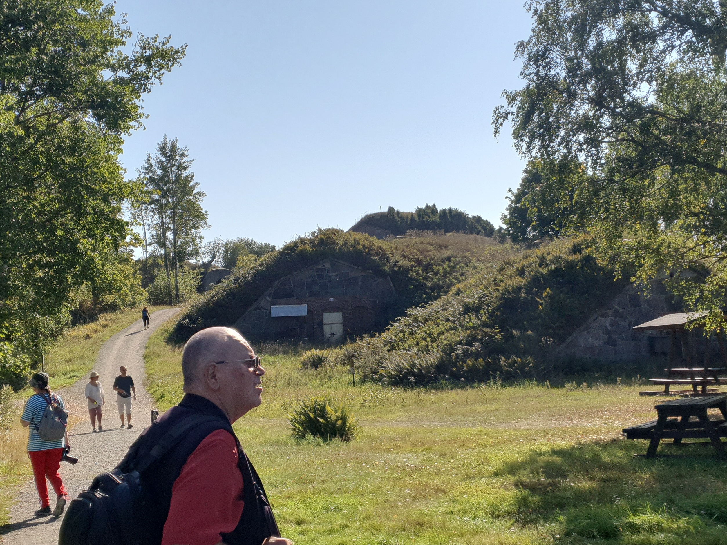 Näkymä Vallisaaren linnoituksilta saaren luoteisrannikolla. Maastoon on rakennettu kiviseinäisiä ruutivarastoja, jotka venäläiset rakensivat 1860-1870-luvuilla. Ne on katettu maakerroksella, ja ne saavat maaston kumpuilemaan. Hiekkainen kävelytie kiemurtelee kumpujen välistä. Vasemmalla näkyy holvikaarimainen betoninen sisäänkäynti varastoon. rephoto