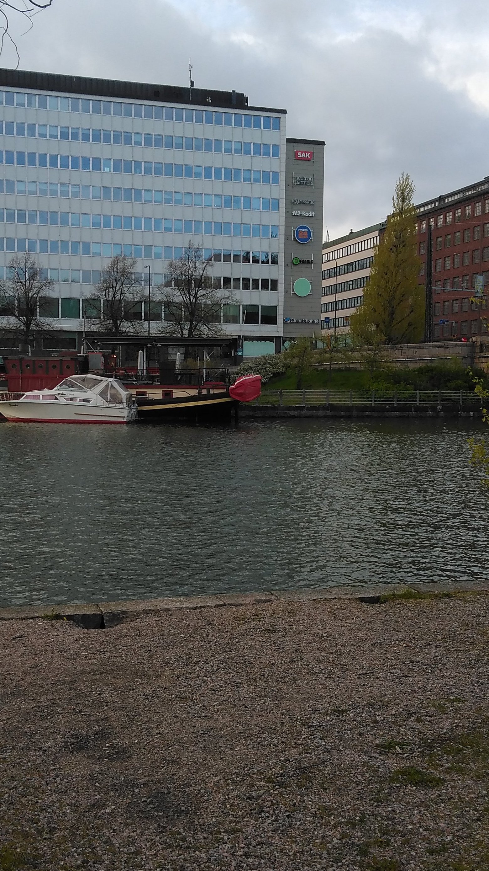 Kaksi henkilöä nostamassa venettä Kaisaniemenlahden rannalla. Etualalla kaksi lasta ja koira. Taustalla Pitkänsillanranta 3 ja Siltasaarenkatu 1. rephoto