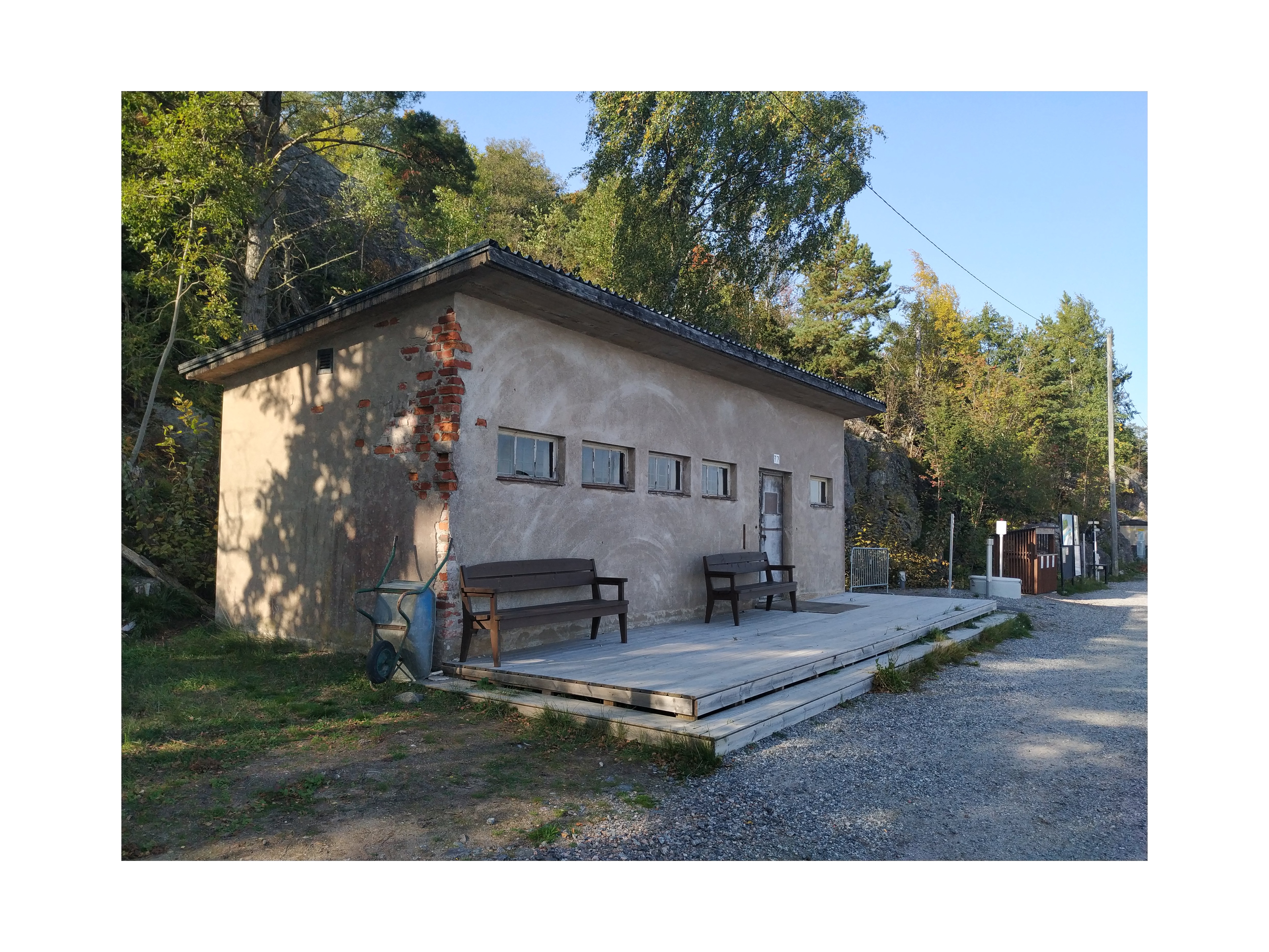 Näkymä Vallisaaren itärantaa kulkevalle tielle. Se on retkisataman tuntumassa jo suurin piirtein Kuninkaansaarta vastapäätä. Punatiilinen rakennus vasemmalla liittyy saaren torpedoluolaston toimintaan. Rakennus on rapattu harmaalla laastilla, josta osa on karissut pois. Rakennuksen päätyä vasten nojaa lavoja ja julkisivun edustalle on rakennettu alusta. Äärimäisenä oikealla pilkottaa meri. rephoto