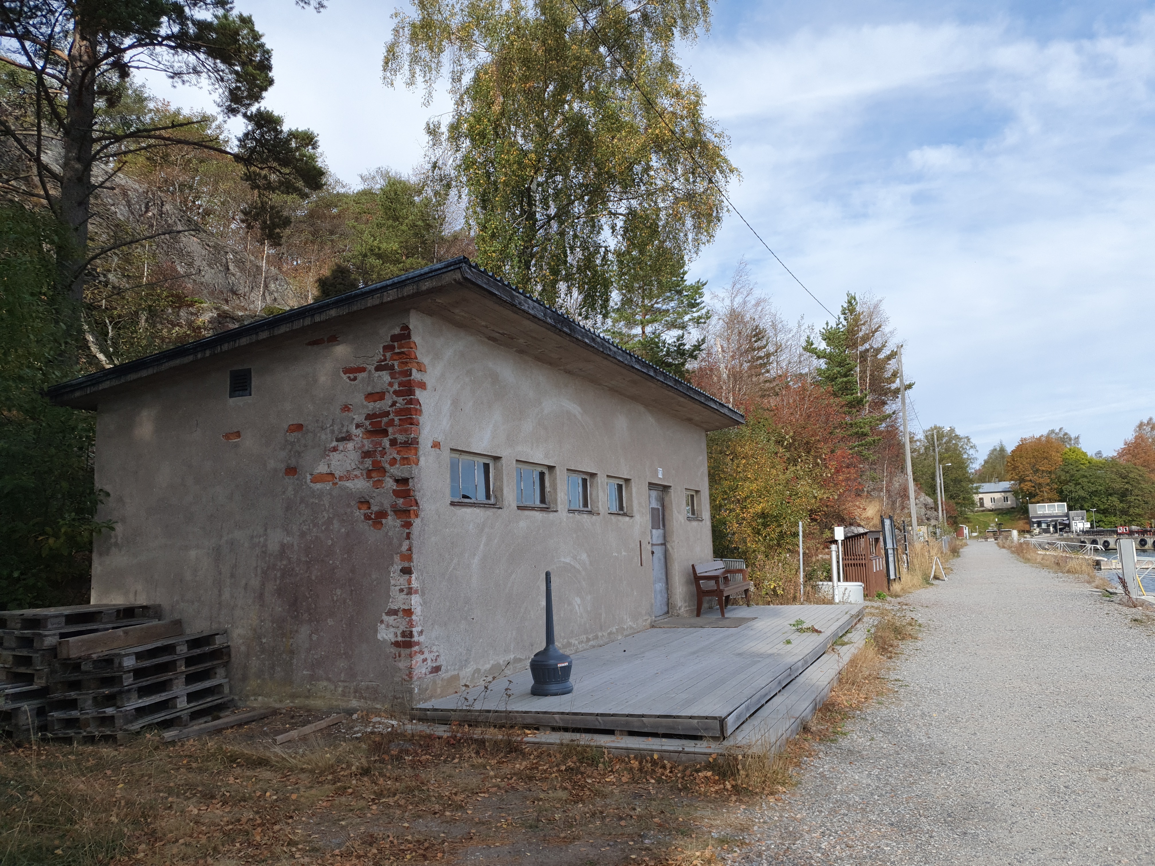 Näkymä Vallisaaren itärantaa kulkevalle tielle. Se on retkisataman tuntumassa jo suurin piirtein Kuninkaansaarta vastapäätä. Punatiilinen rakennus vasemmalla liittyy saaren torpedoluolaston toimintaan. Rakennus on rapattu harmaalla laastilla, josta osa on karissut pois. Rakennuksen päätyä vasten nojaa lavoja ja julkisivun edustalle on rakennettu alusta. Äärimäisenä oikealla pilkottaa meri. rephoto