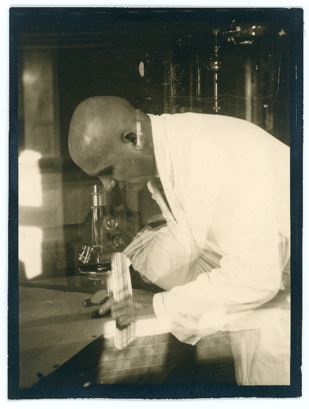 Johannes Vares valges kitlis kummardamas mikroskoobi kohale. Ülepildistatud foto?