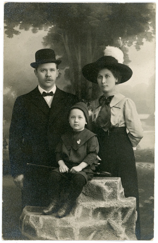 Portreefoto kaabuga mehest, laia äärega kübaraga naisest ja istuvast lapsest.