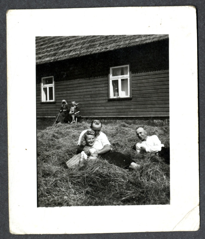 Osvald Asi, tundmatu naine ja mees lamavad elumaja ees heinakuhjas. Maja seina ääres istub vanem naisterahvas koos kahe väikese lapsega
