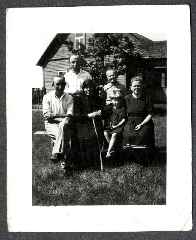 Osvald Asi perekonnafoto: mees, musta rõivastatud vanem naine kepiga, tüdruklaps ja noor naisterahvas istuvad pingil, meesterahvas ja poisslaps seisavad nende taga. Taustal elumaja