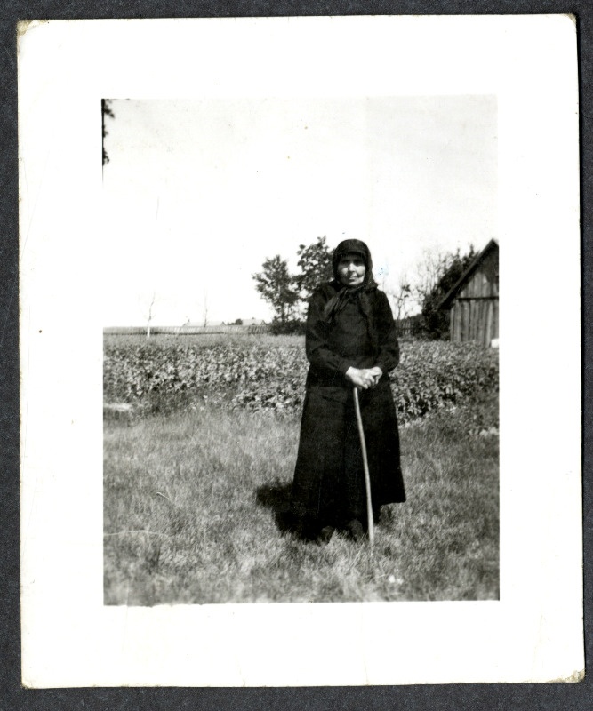 Musta rõivastatud pearätikuga vanem naine seisab kepile toetudes peenra või põllu ääres. Eemalt paistab taluhoone nurk, puud ja aed