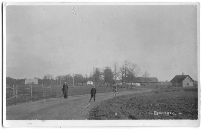 Külatänav Emmastes.Teel kaks jalakäijat, üks jalgrattur, tagaplaanil koorejaam, endine mõisa viinavabrik