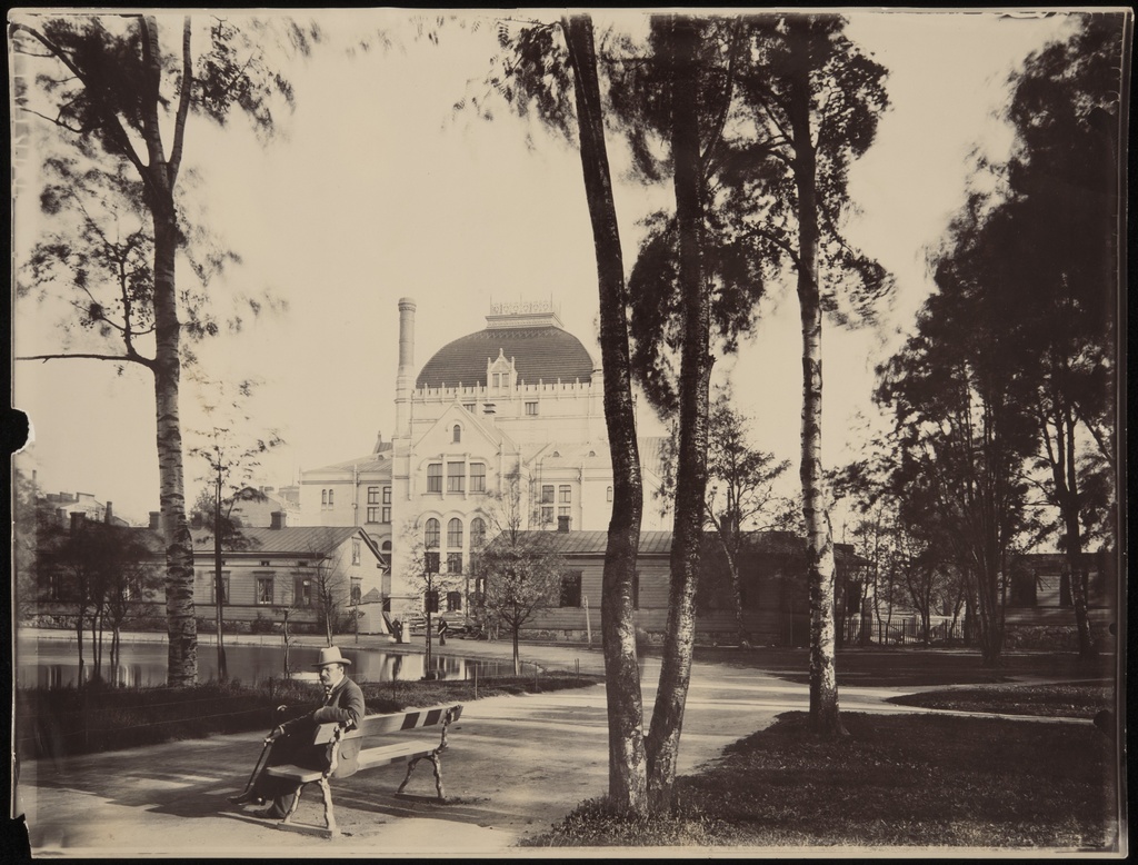 Kaisaniemen puisto, mies istuu penkillä. Vasemmalla joutsenlampi, taustalla Kansallisteatteri ja Villensaunan (Wilhelmsbad) kylpylän puurakennuksia.