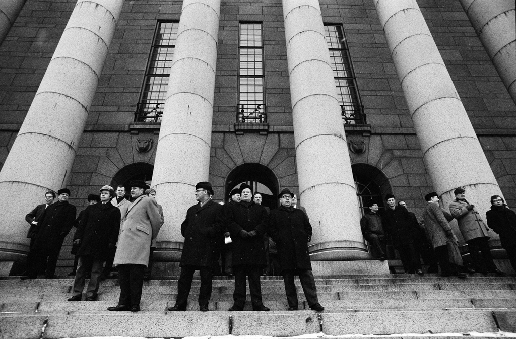 Miehiä seuraamassa Eduskuntatalon portailla pylväiden edustalla korkeakouludemokratiaa vaativaa mielenosoitusta.