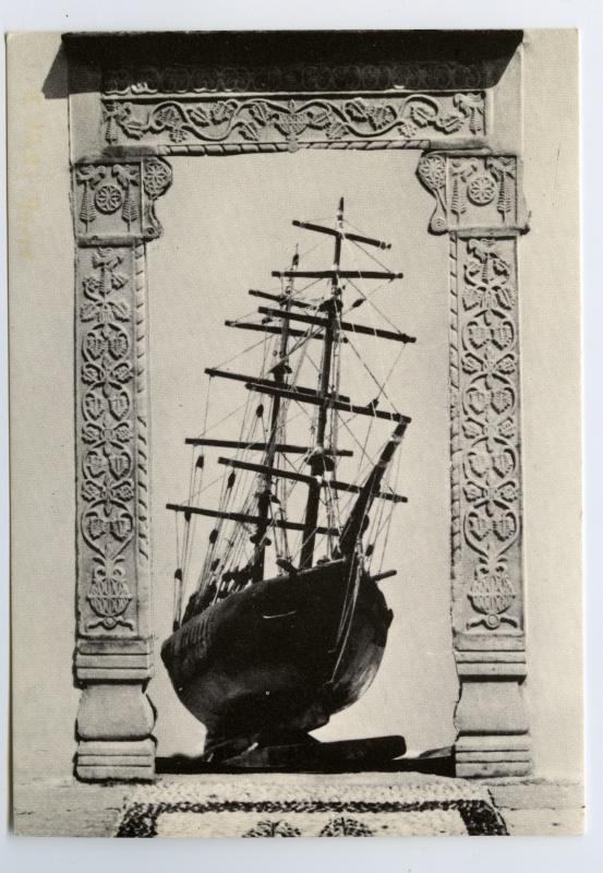 Mykonose Etnograafiamuuseumi postkaart: taimornamentidega ukseraam ja selles 2-mastilise purjelaeva mudel