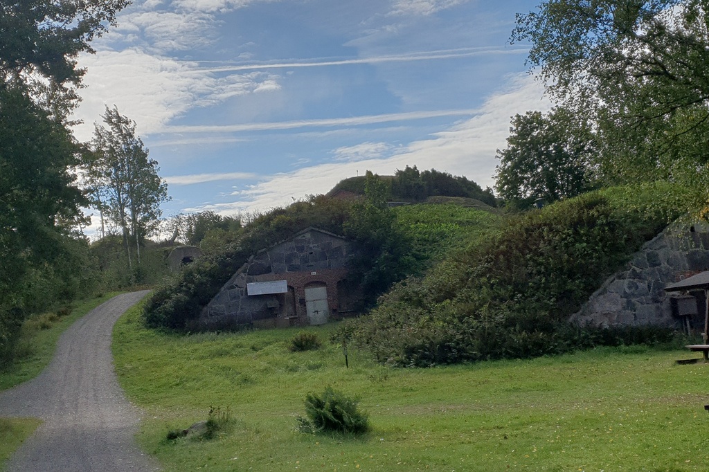 Näkymä Vallisaaren linnoituksilta saaren luoteisrannikolla. Maastoon on rakennettu kiviseinäisiä ruutivarastoja, jotka venäläiset rakensivat 1860-1870-luvuilla. Ne on katettu maakerroksella, ja ne saavat maaston kumpuilemaan. Hiekkainen kävelytie kiemurtelee kumpujen välistä. Vasemmalla näkyy holvikaarimainen betoninen sisäänkäynti varastoon. rephoto