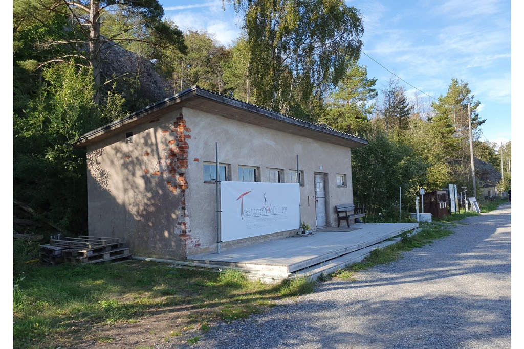 Näkymä Vallisaaren itärantaa kulkevalle tielle. Se on retkisataman tuntumassa jo suurin piirtein Kuninkaansaarta vastapäätä. Punatiilinen rakennus vasemmalla liittyy saaren torpedoluolaston toimintaan. Rakennus on rapattu harmaalla laastilla, josta osa on karissut pois. Rakennuksen päätyä vasten nojaa lavoja ja julkisivun edustalle on rakennettu alusta. Äärimäisenä oikealla pilkottaa meri. rephoto