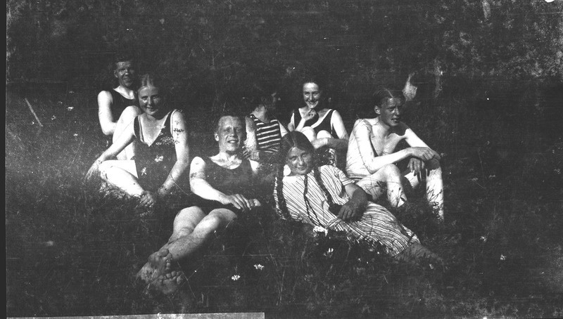 Seitse inimest istumas-pikutamas rannal. Ees patsidega triibulises kleidis tütarlaps.