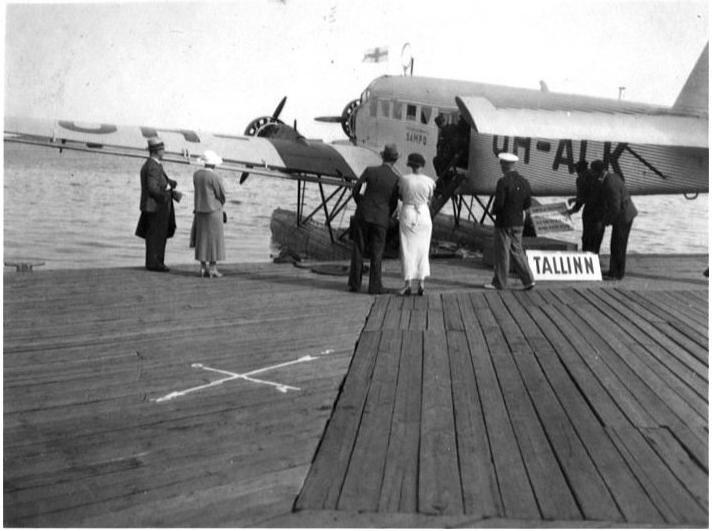 Vesilentokone lentosataman laiturissa. Muutamia ihmisiä katselee laiturissa olevaa matkustajalentokonetta, joka on todennäköisesti mallia Junkers Ju 52. Koneen kyljessä näkyy nimi "Sampo", ja sen tunnus on OH-ALK. Koneen ohjaamon yläpuolella liehuu pieni