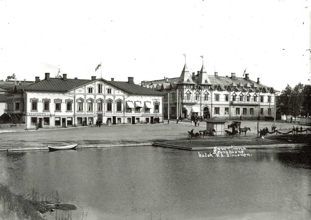 Miljonatalon rannasta yli Haapasalmen itään. Sikion talossa Suomen lippu, "Kelloseppä", "Kukka- ja siemenkauppa", "Kangaskauppa", "Sekatavarakauppa", "Ruokatavarakauppa", "Kangastavarakauppa" (Huttunen & Hirvonen), "Pankki". Seurahuone liputettu Suomen lipuin. kesä 1923. View east from Miljoonatalo