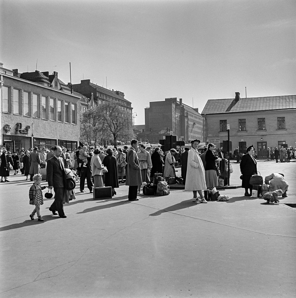 Matkustajia Linja-autoasemalla Lasipalatsin sisäpihalla. Valkoiseen takkiin pukeutunut nainen todennäköisesti Maire Ekholm.