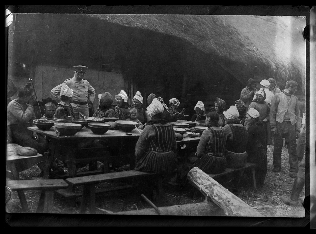 Big party in Muhumaa. A number of women in clothes sits at the table, big bowls in front. One woman talks with a mundrier who stands in the hip. Men stand away and talk to each other (repro)