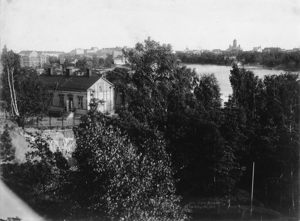 Rautatieläisten asuinrakennus Eläintarhanlahden rantatienoolla. Talo on kallioleikkauksen reunalla, ja alhaalla puiden takana kulkee rautatie.