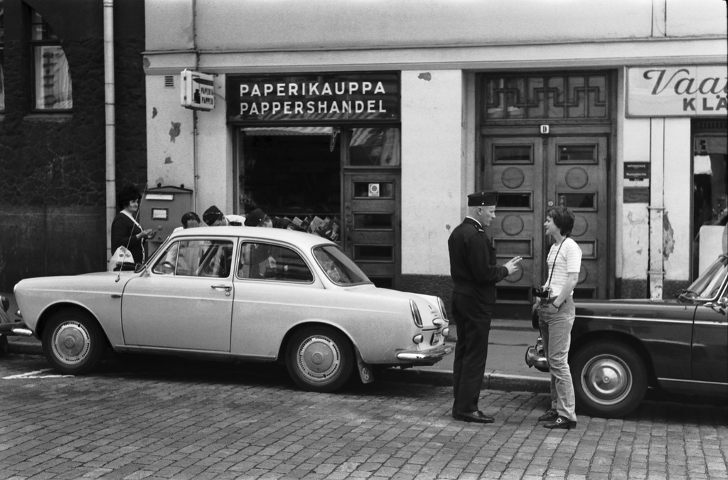 Liisankatu 18. Pari poliisia auttamassa ihmisiä vaalean auton oven avaamisessa pienen Paperikaupan edustalla. Yksi poliiseista puhuttelemassa valokuvaajaa Eeva Ristaa tapahtuman valokuvaamisesta.