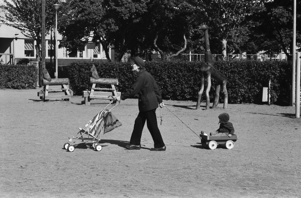 Tehtaanpuisto. Lastenrattaita työntävä iäkäs nainen vetämässä lasta pienissä kärryissä Tehtaanpuiston leikkipaikalla. Taustalla suuria puueläimiä.