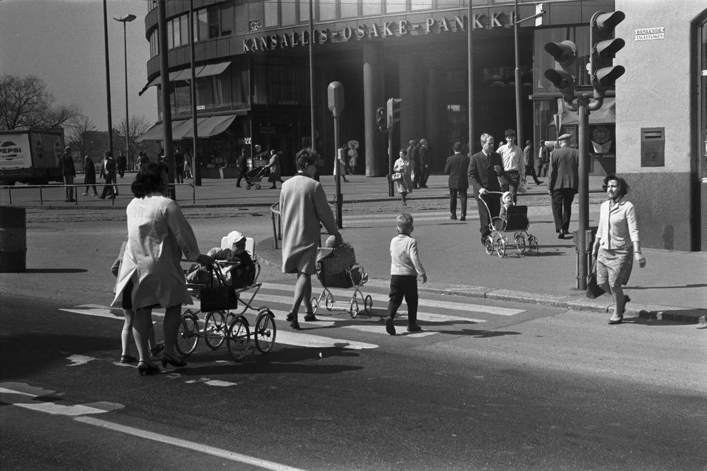 Hämeentie. Kaksi naista lasten ja rattaiden kanssa ylittämässä katua Hakaniementorin kulmalla Hämeentien ja Siltasaarenkadun risteyksessä. Taustalla Ympyrätalo.
