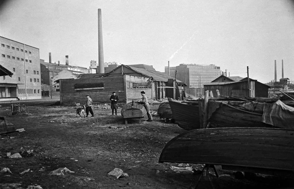 Veneitä, ränsistyneitä puurakennuksia ja miehiä Sörnäisten rannassa. Vasemmalla Sörnäisten rantatie 19.