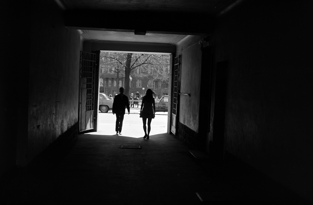 Mies ja nainen kävelemässä Agricolankatu 13:n porttikäytävässä. Nainen kävelemässä ulos kadulle, mies tulossa kohti sisäpihaa. Taustalla Karhupuisto.