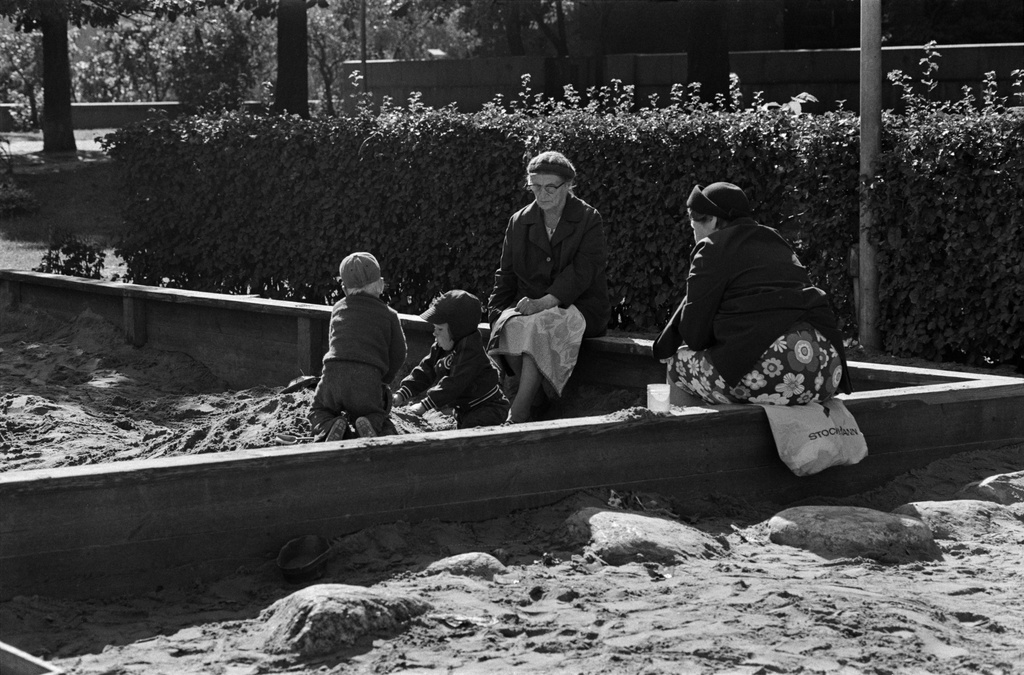 Tehtaanpuisto. Kaksi lasta leikkimässä hiekkalaatikossa Tehtaanpuiston leikkipaikalla. Laatikon reunalla istumassa kaksi iäkästä naista.