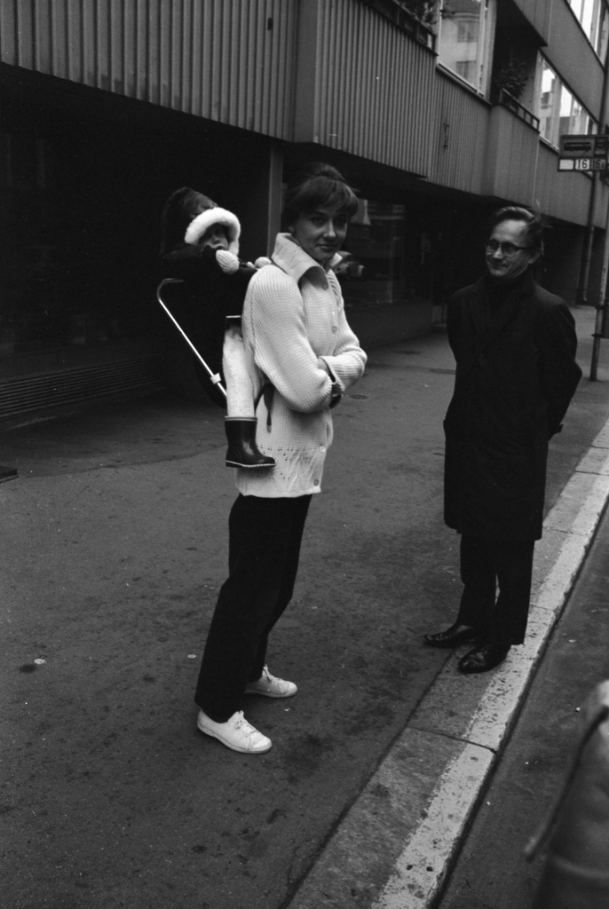 Punavuori. Mies ja nainen kantotelineessä oleva lapsi selässään seisomassa Perämiehenkatu 5:n edustalla. Taustalla linja-autopysäkki.