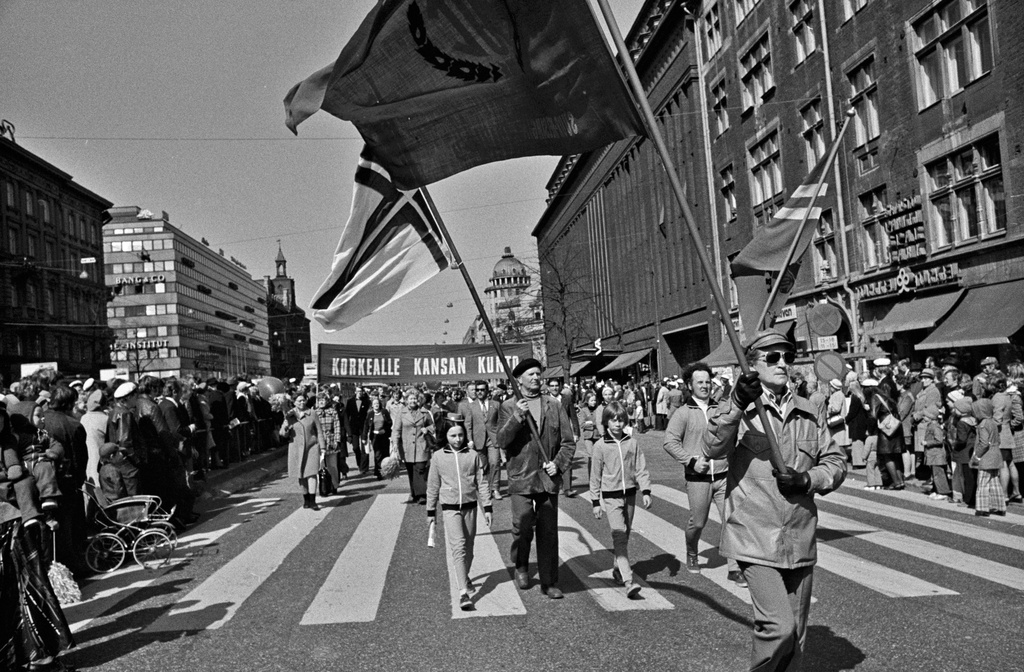 Vappumarssijoita Mannerheimintiellä Stockmannin tavaratalon kohdalla. Kulkueen edessä lipunkantajia, takana banderolli, jossa teksti: Korkealle kansan kunto. Yleisö seuraa marssia kadun varrella.