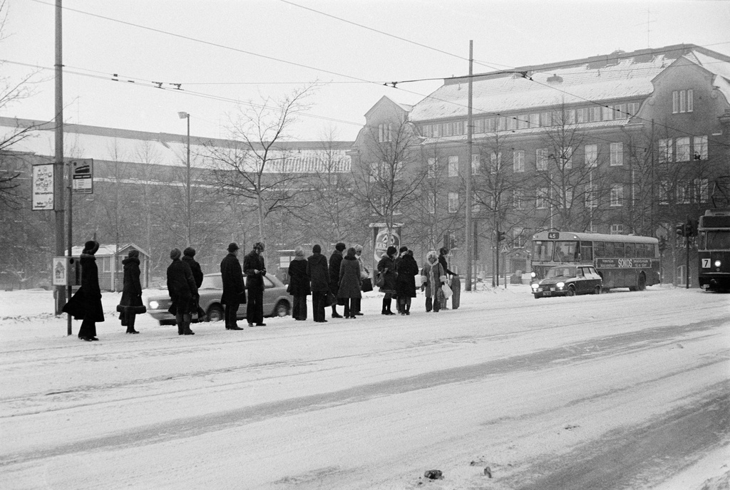 Ensilumi Töölössä vuonna 1973. Liikennettä Runeberginkadulla. Ihmisiä odottamassa raitiovaunua raitiovaunupysäkillä. Taustalla näkyy entinen Tyttönormaalilyseo, vuodesta 1969 Kauppakorkeakoulun Chydenia.