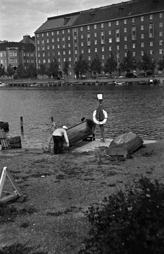 Kolme henkilöä nostaa venettä Kaisaniemenlahden rannalla. Taustalla Pitkänsillanranta 7 - 13.