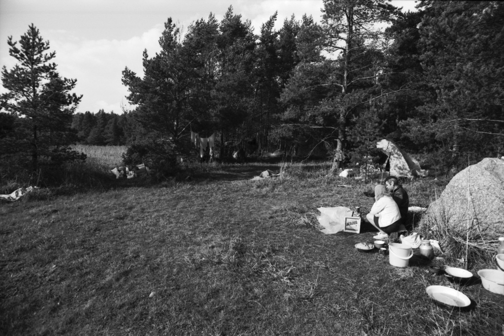 Kallahdenniemi. Mies ja nainen ruokailemassa Kallahdenniemen rannassa lähellä telttaa, jossa vielä syksyllä asustavat.Taaempana puiden välissä pyykkiä kuivumassa.