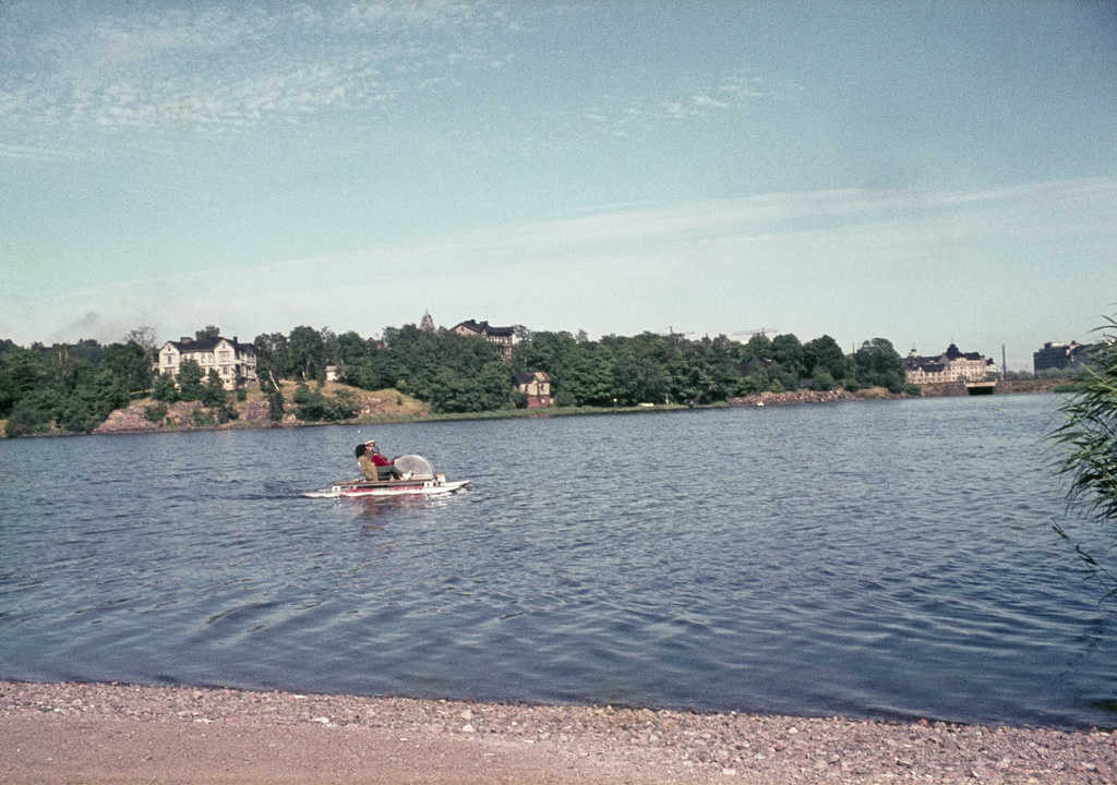 Töölönlahden rantaa, vedessä perhe polkuveneellä, taustalla Linnunlaulun huviloita.
