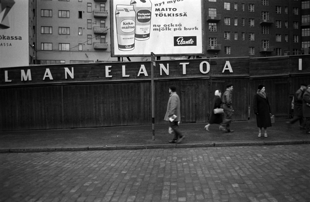 Hämeentie 16. Ihmisiä bussipysäkillä ja jalankulkijoita kadulla. Taustalla lauta-aita ja Elannon tölkissä myytävän maidon mainos. Kuva vuodelta 1958, jolloin maitotölkit tulivat käyttöön. Hämeentie 16 tyhjä tontti. Sen takana Neljäs linja 3.