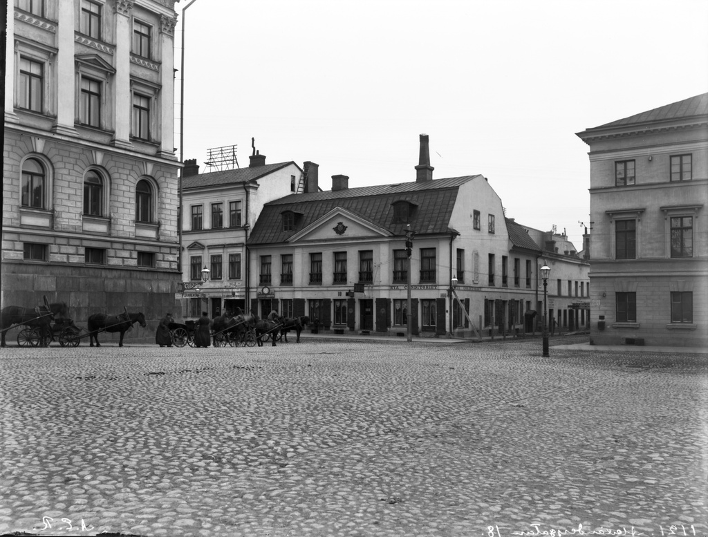 Aleksanterinkatu 18 (- Katariinankatu 3) nähtynä Senaatintorilta, vasemmalla Senaatintalo (=Valtioneuvoston linna, Nikolainkatu 1, nyk. Snellmaninkatu 1) ja vuokra-ajuriasema