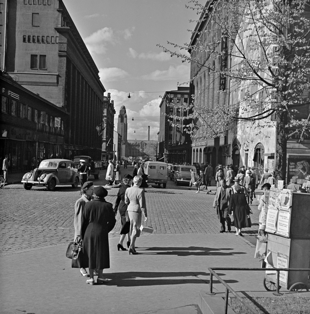 Jalankulkijoita Pohjoisesplanadin ja Keskuskadun risteyksessä. Taustalla Keskuskatu ja rautatieasema. Oikealla lehtien myyntikoju. Vasemmalle kääntyvä auto on Ford vm. 1937.