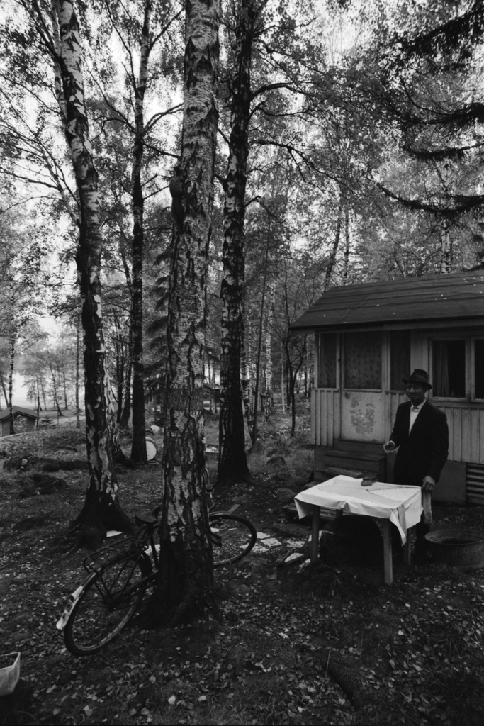 Kulosaari. Näkymä syksyiseltä Kivinokan kesämaja-alueelta. Mökin pihalla mies pesemässä juuriharjalla valkoisella vahakankaalla peitetyn pöydän päälle levitettyjä pitkiä alushousuja. Koivunrunkoa vasten nojaa miesten polkupyörä.