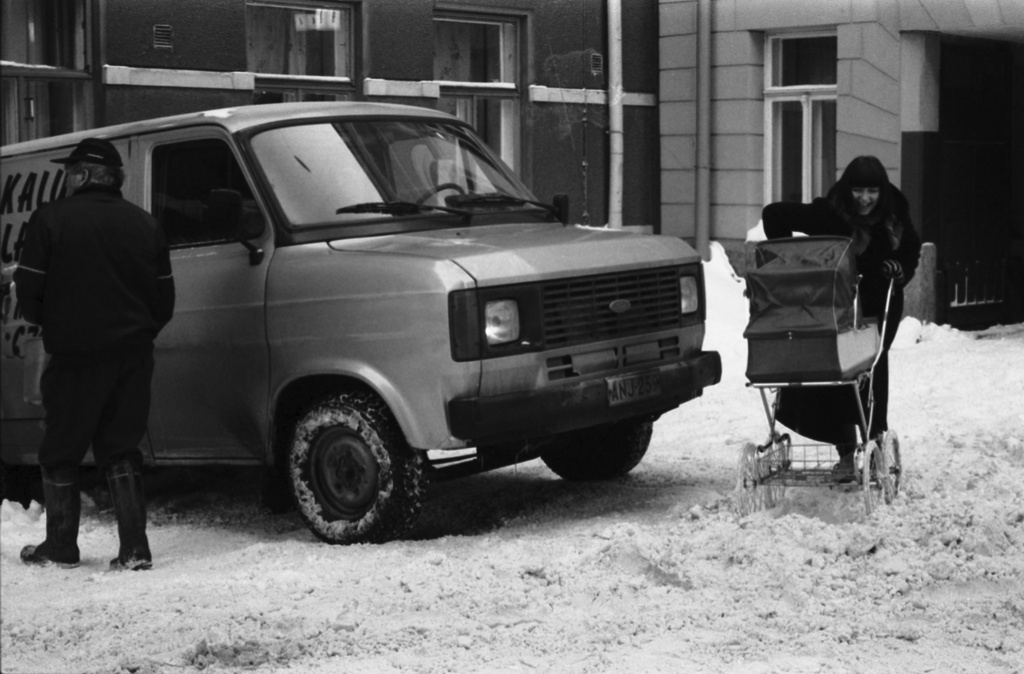 Kulmakatu. Äiti (Etta Harvilahti) 1970-luvulta peräisin olevien muovisten, kapeapyöräisten lastenvaunujen kanssa ohittamassa Kulmakatu 4a:n edustalle jalkakäytävälle lumikasaan juuttunutta pakettiautoa.
